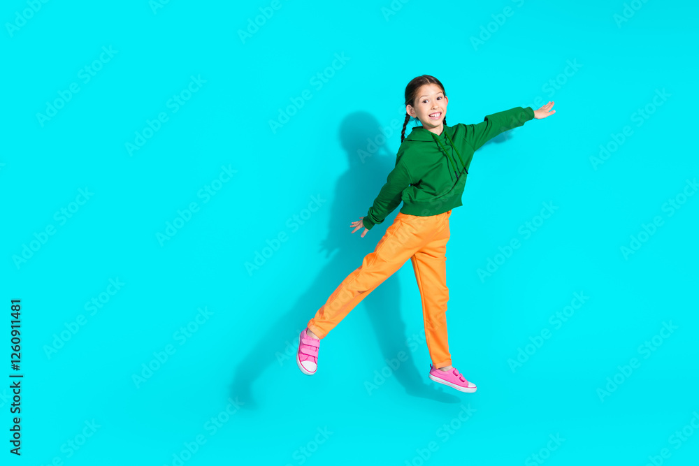 Obraz premium Adorable young girl in orange pants and green hoodie smiling brightly against teal background