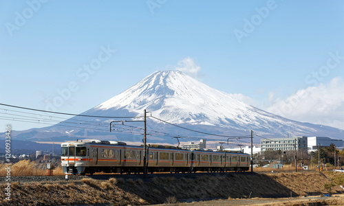 A local train of JR Gotemba Line (御殿場線) traveling through the countryside on a sunny winter day and snow capped Mount Fuji dominating the background under blue clear sky in Susono, Shizuoka, Japan photo