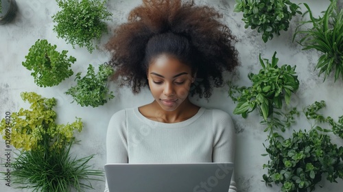 Young woman working on laptop surrounded by green plants in a bright indoor s...
