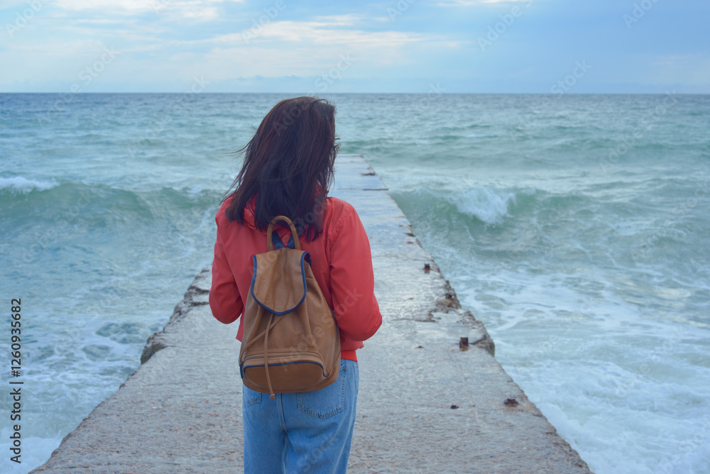 Rear view of a young woman tourist with a backpack standing on the pier and waves hitting the pier in a storm. City of Sudak, Crimea.