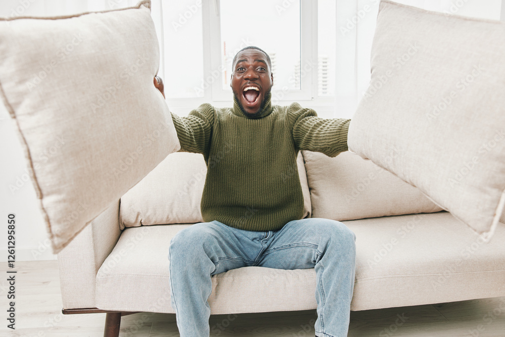 Happy Black man playfully holding pillows while sitting on a light colored couch in a bright, airy room, showcasing joy and relaxation Happy Black man playfully holding pillows while sitting on a light colored couch in a bright, airy room, showcasing joy and relaxation