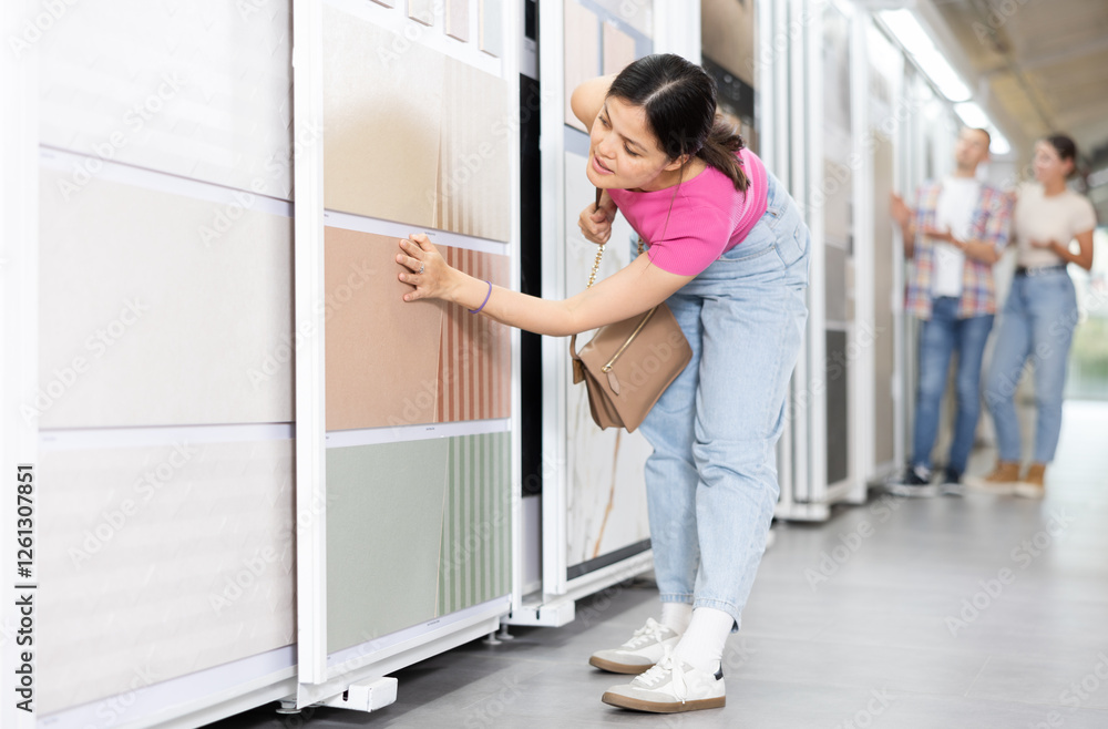 Positive young Asian woman selecting ceramic tiles for home renovation in building hypermarket, carefully examining options presented on sliding display panels Positive young Asian woman selecting ceramic tiles for home renovation in building hypermarket, carefully examining options presented on sliding display panels