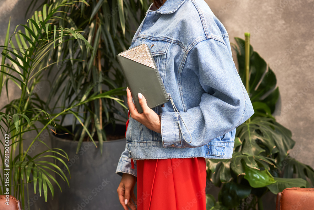 Fashionable woman in denim jacket holding stylish diary in urban greenery setting, modern lifestyle concept
