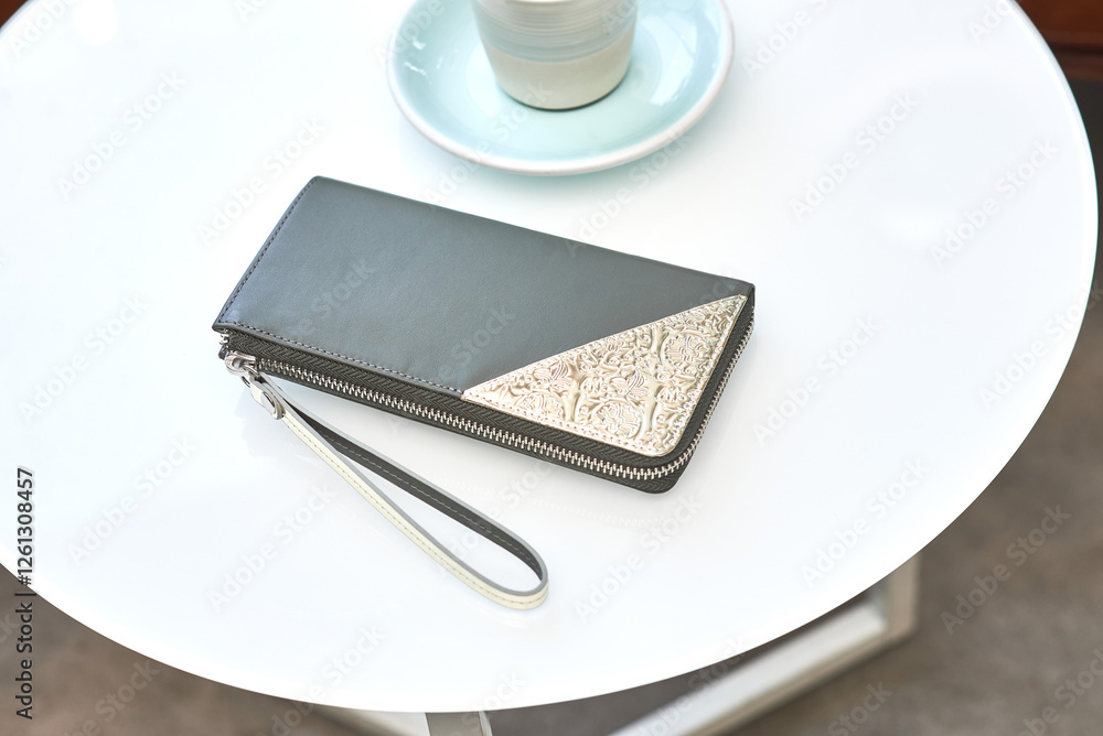 Elegant leather wallet with intricate metal detail on white table, next to cup and saucer, fashion accessory style concept
