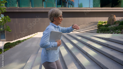 Caucasian mature business woman hurries walking up stairs office building in city looking at hand watch busy senior adult businesswoman female employer running fast to work with folder lateness time photo