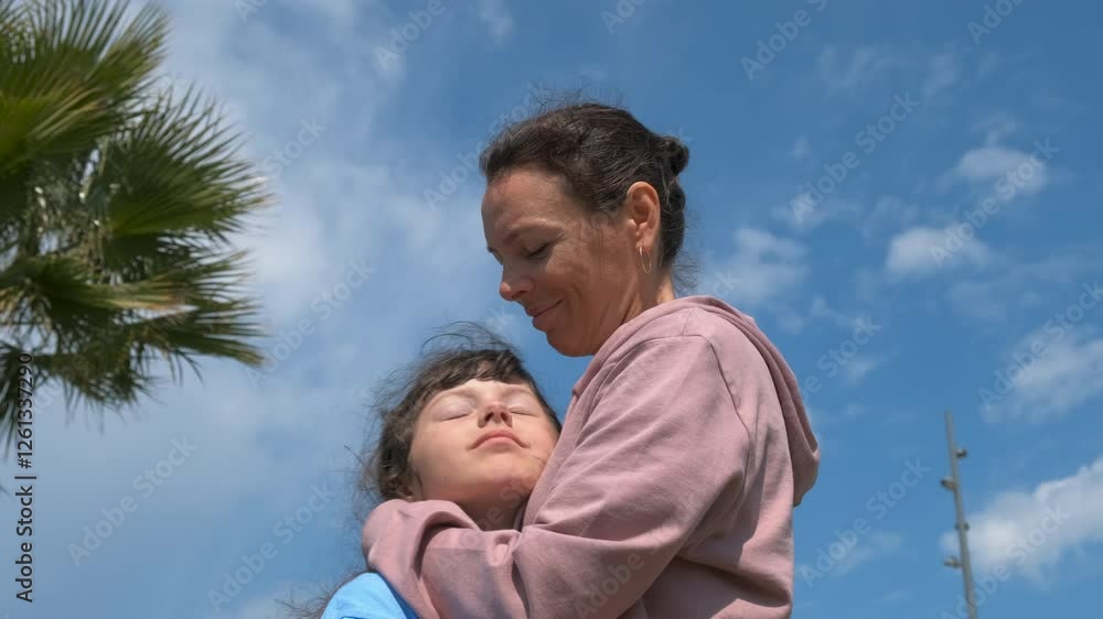 Family hugs against palm tree. A mother with love embrace her daughter against palm trees in the garden. A concept of celebrating mother's day with parents in summer.