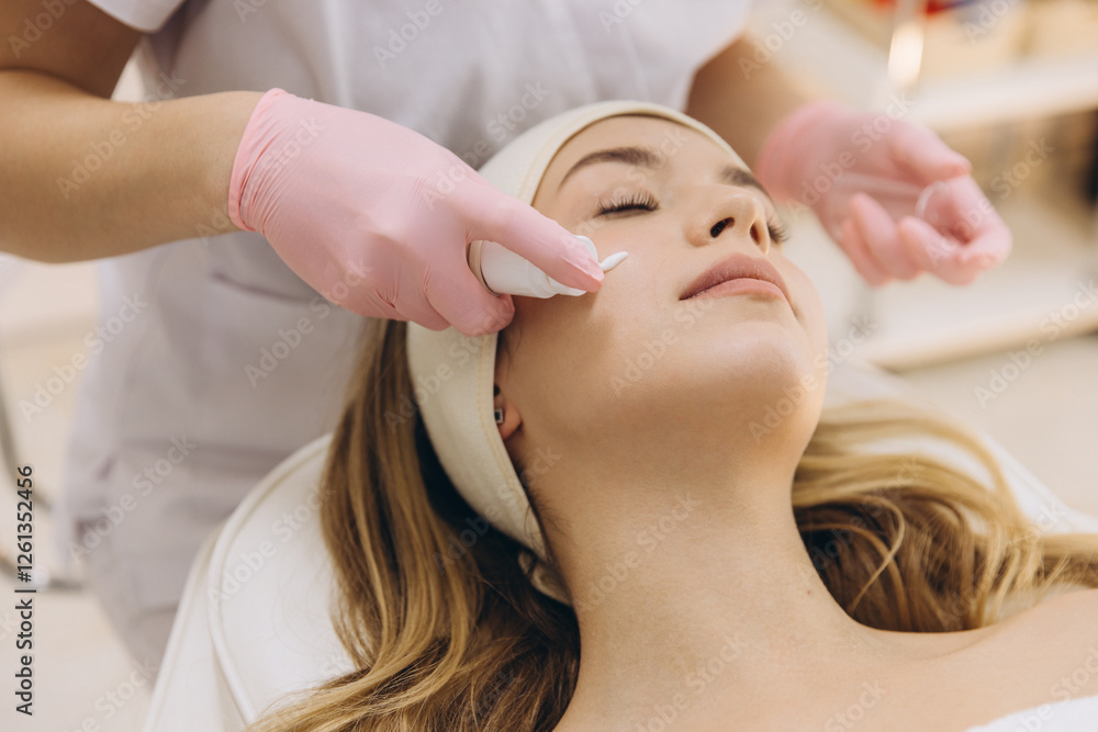 Naklejka premium Close up of cosmetologist applying cream on female face during skin care procedure in beauty salon