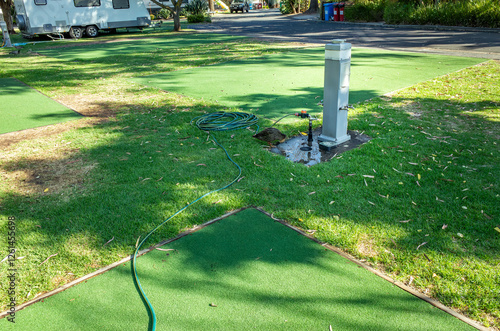 Canvastavla A water service point stands on an artificial green turf surface at a caravan park, with a water hose coiled nearby for camper access