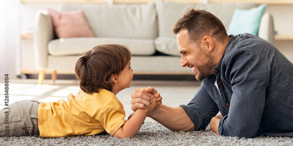 Bringing up strong kid. Happy father competing arm wrestling match with his little boy, lying together on floor at home and smiling to each other, profile portrait Bringing up strong kid. Happy father competing arm wrestling match with his little boy, lying together on floor at home and smiling to each other, profile portrait