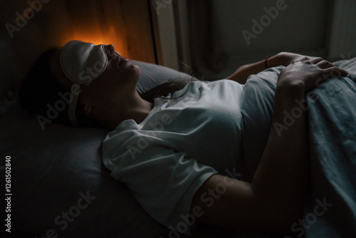 Fotografie A woman wearing a sleep mask is resting in bed, lying under a cozy blanket in a dimly lit room
