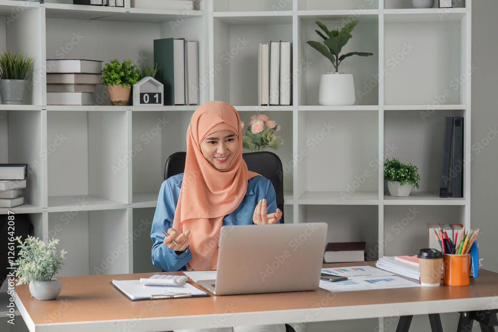 Muslim woman happily discussing in video call while working on a laptop at her desk. Muslim woman happily discussing in video call while working on a laptop at her desk.