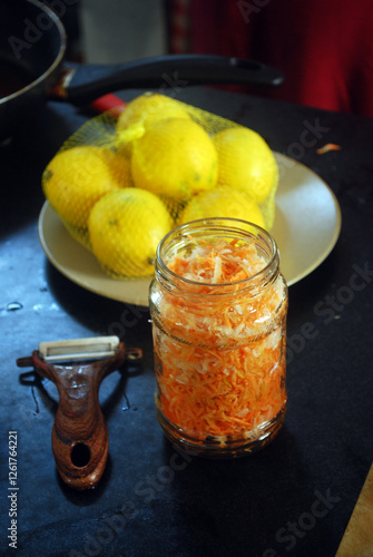 A jar of Hokben salad. Made of pickled carrots and white radish sits on a dark kitchen counter, accompanied by a peeler, and a plate of lemons in a net bag. Selective focus photo