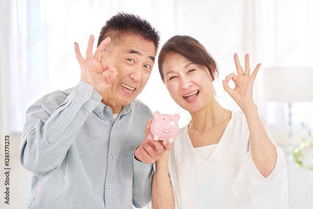 Φωτογραφία 貯金箱を持つシニア夫婦　OKサイン　リビング