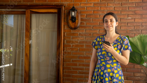 Obraz na plátně Modern Lifestyle: A portrait of a smiling woman holding a phone, she is standing against a brick wall, embodying contemporary life