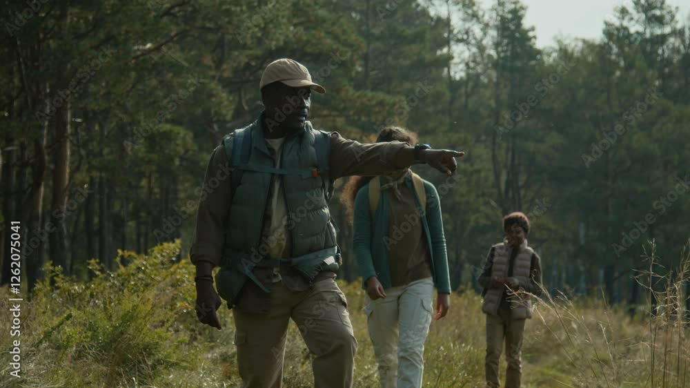 African American dad pointing at lake when slowly walking down path with two kids following him during weekend hike African American dad pointing at lake when slowly walking down path with two kids following him during weekend hike