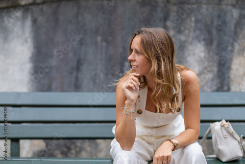 Stylish woman in white outfit sitting thoughtfully on a bench