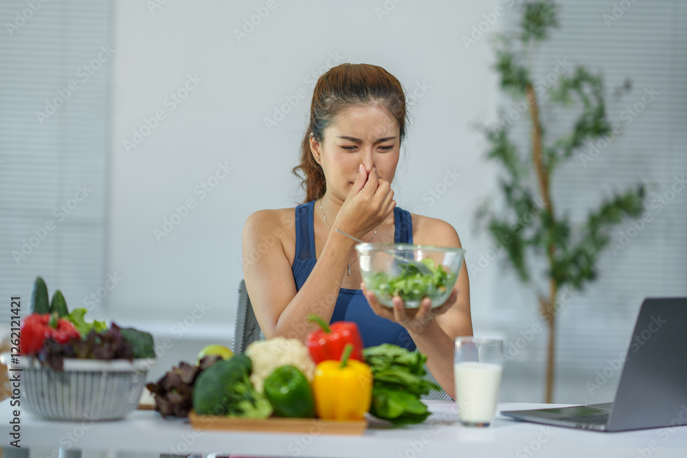 Young asian woman pinching her nose due to bad smell coming from a bowl of salad, sitting at the table with many vegetables and a glass of milk, having breakfast at home Young asian woman pinching her nose due to bad smell coming from a bowl of salad, sitting at the table with many vegetables and a glass of milk, having breakfast at home