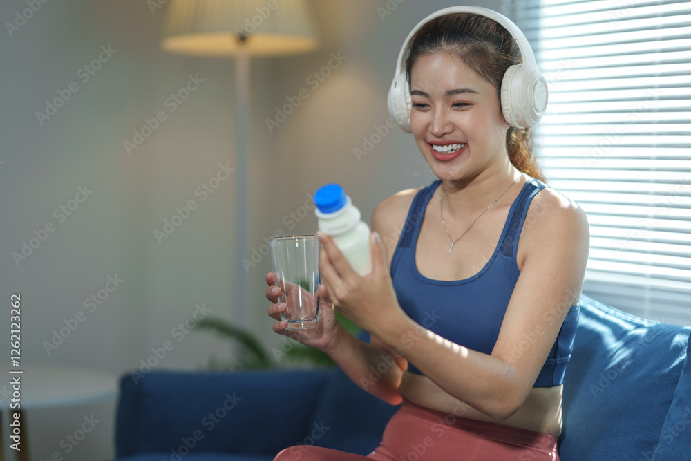 Young woman in workout attire enjoying music with headphones, holding a glass and a beverage bottle, sitting on a comfortable sofa in a cozy living room setting Young woman in workout attire enjoying music with headphones, holding a glass and a beverage bottle, sitting on a comfortable sofa in a cozy living room setting
