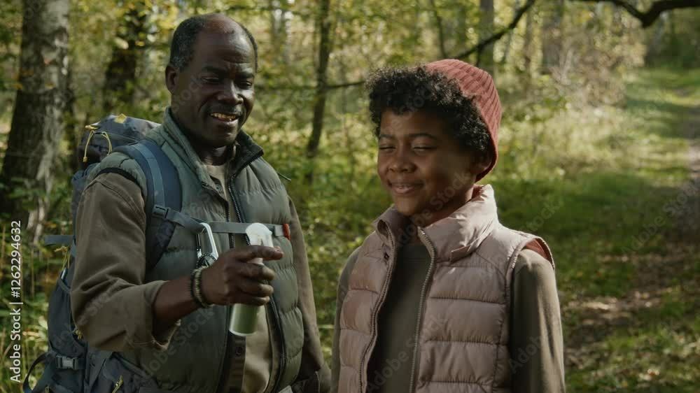 Tilt up view of positive dad with backpack spraying mosquito repellent on sweater and vest of smiling boy in woods Tilt up view of positive dad with backpack spraying mosquito repellent on sweater and vest of smiling boy in woods