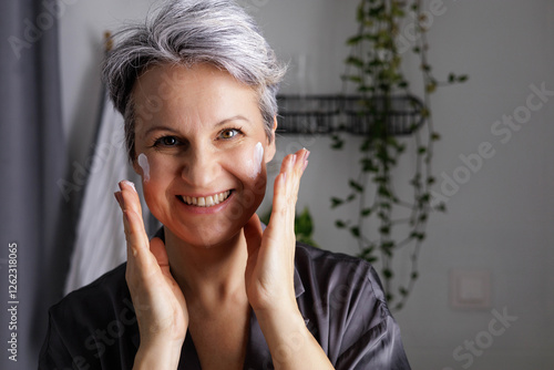 Mature beautiful woman during menopause with gray hair applies hormonal anti-aging cream to her face