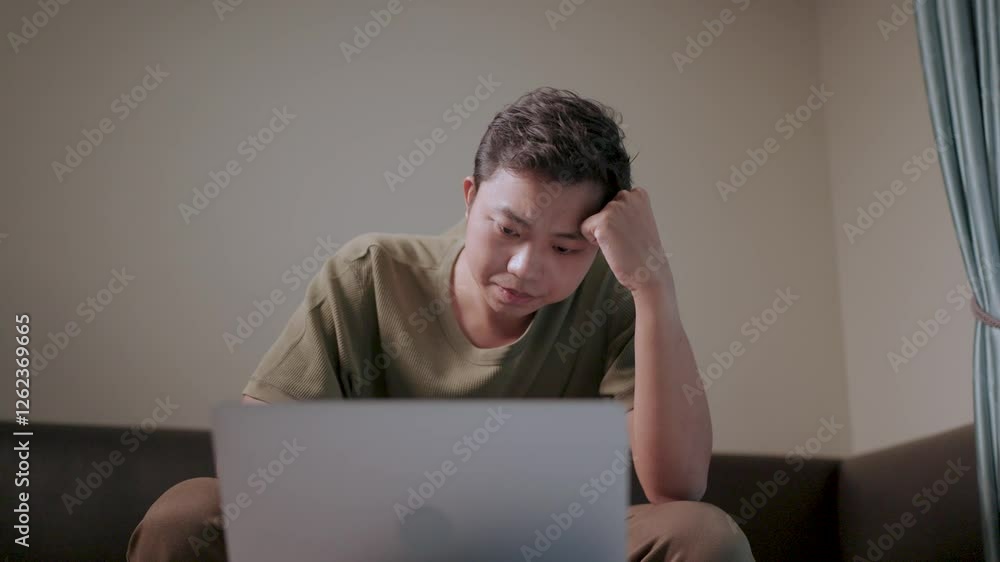 Focused young man working on laptop. A young man sits on a couch, deeply engrossed in his laptop, his hand resting thoughtfully on his forehead.
