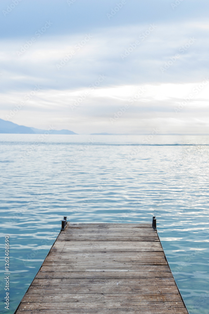 Naklejka premium Tranquil scene featuring a wooden pier extending into a calm blue lake, with distant mountains and a cloudy sky creating a peaceful atmosphere.
