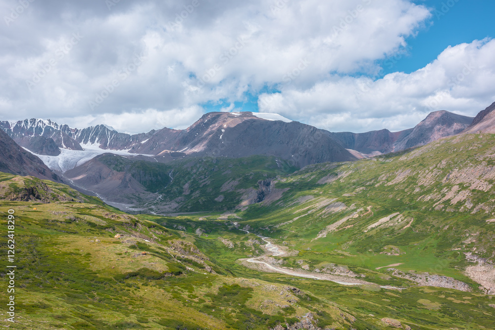 Wide green alpine valley with snake river among hills and rocks with vast view to large snow mountain top and big glacier tongue under cloudy sky. Serpentine river in high mountains. Shadows of clouds Wide green alpine valley with snake river among hills and rocks with vast view to large snow mountain top and big glacier tongue under cloudy sky. Serpentine river in high mountains. Shadows of clouds
