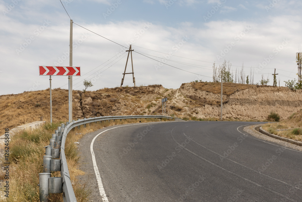 A curved road navigates through a dry landscape in Tajikistan, with sparse vegetation and a cloudy sky above, showcasing the region's rugged terrain. A curved road navigates through a dry landscape in Tajikistan, with sparse vegetation and a cloudy sky above, showcasing the region's rugged terrain.