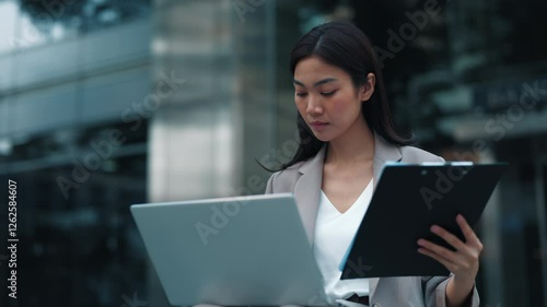 Portrait of Thai woman entering data from work papers into laptop sitting on city street.