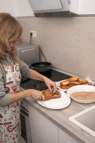 Woman wearing a festive apron placing freshly made torrijas on a plate after coating them in cinnamon sugar. photo