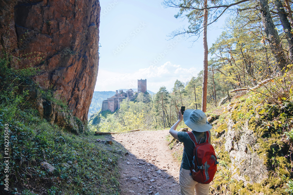 Obraz premium A woman wearing a sun hat and a red backpack takes a photo of The Three Castles of Eguisheim while hiking through a scenic forest trail with rugged cliffs and lush greenery.