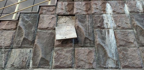 Wall of the building. Textured wall covered with stone tiles. The tiles are uneven and imitate real stone. One tile has fallen off and is hanging on a metal fastener cement wall is visible behind it.