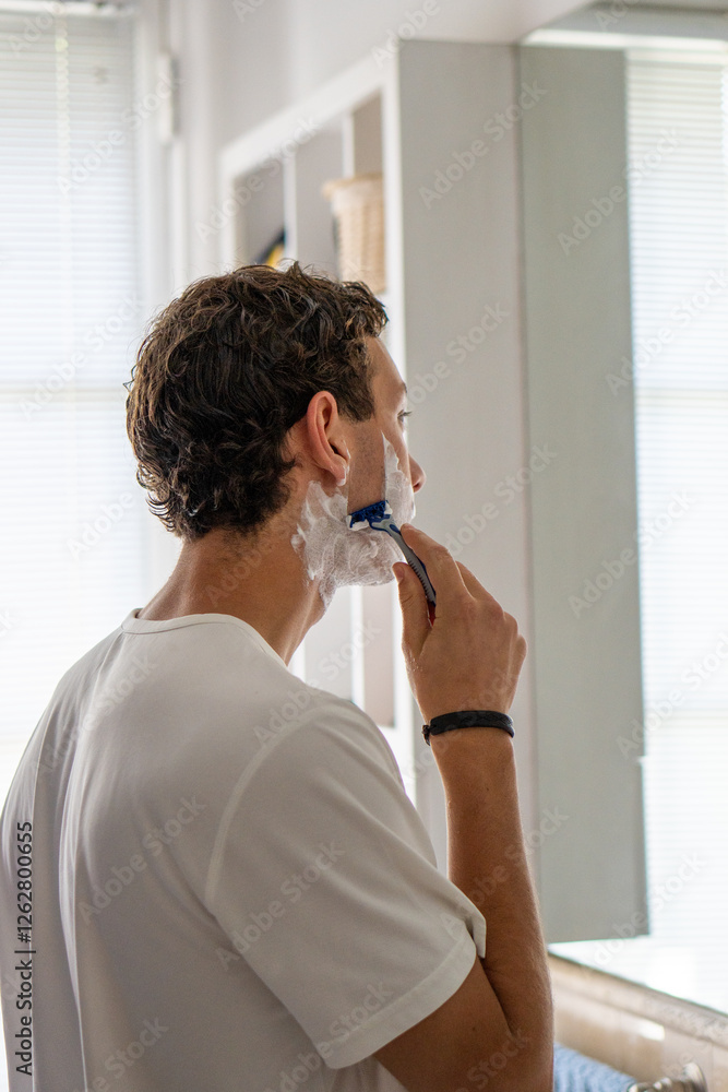 A vertical image of a young man carefully shaving his beard while standing in front of a bathroom mirror, focusing on personal grooming and daily routines.