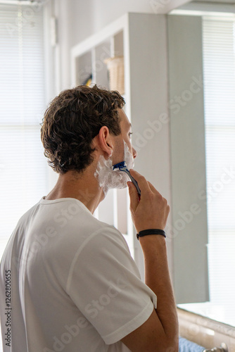 A vertical image of a young man carefully shaving his beard while standing in front of a bathroom mirror, focusing on personal grooming and daily routines.