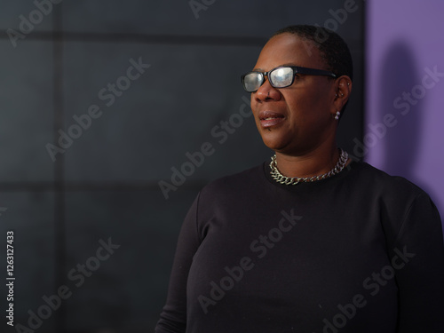 A smiling woman with glasses and a black outfit poses confidently against a modern dark tiled background. Her warm expression conveys positivity and approachability
