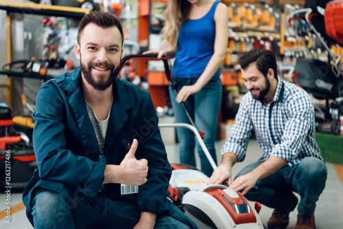 Salesman in blue robe is posing with couple of clients and new lawn mover in power tools store.