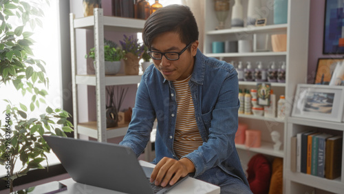 Young man working on laptop in a well-decorated home interior surrounded by various decor items and plants. photo