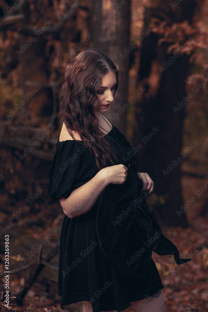 Fototapeta premium This striking photograph features a woman standing in a dark, moody forest, exuding a gothic and mysterious aura. She is dressed in an all-black ensemble, including a sheer, flowing cape.