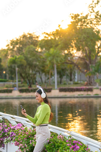 Young Asian Woman Listening to Music with Headphones Using Smartphone in Park Outdoors