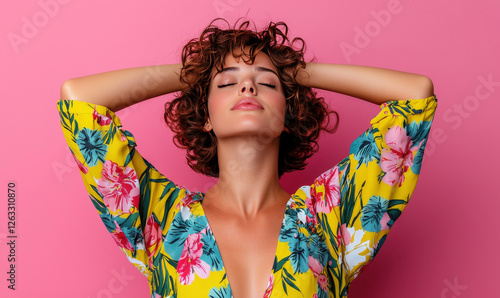 Young woman with curly hair poses gracefully against a vibrant pink background wearing a colorful floral dress, radiating confidence and joy in summer fashion