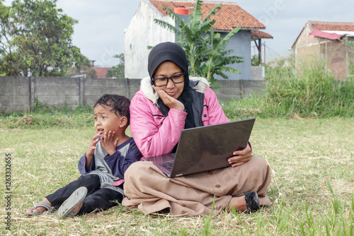 Hijabi Woman Working on Laptop Outdoors with a Child photo