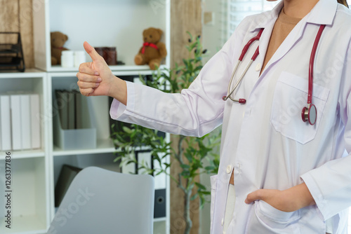 Caucasian pediatrician in hospital examination room. Female doctor in white coat gives thumbs-up gesture, symbolizing approval and encouragement in a professional medical environment. photo