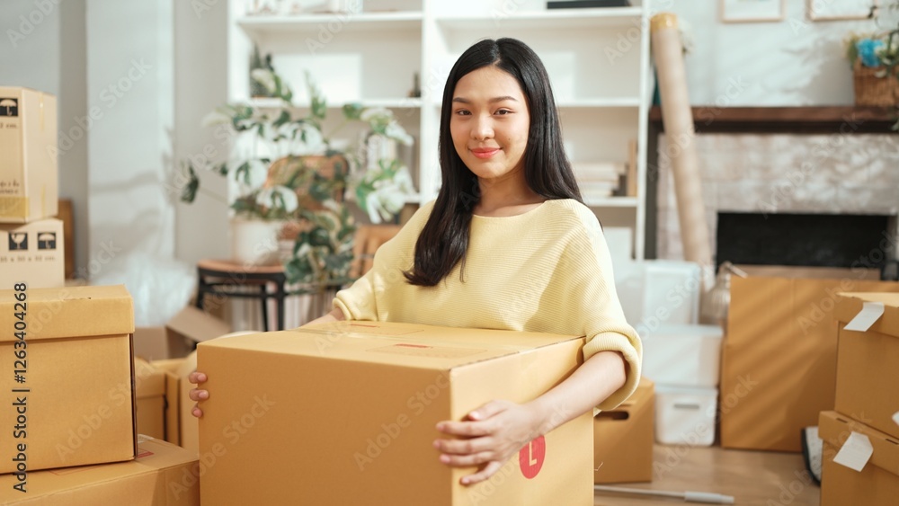 Young female business owner holding cardboard while standing at shop. Attractive happy entrepreneur carrying package and box while preparing for delivery or shipping process at home office. Myrmidon. Young female business owner holding cardboard while standing at shop. Attractive happy entrepreneur carrying package and box while preparing for delivery or shipping process at home office. Myrmidon.