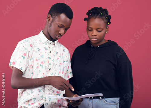 A young man points at a document while a young woman looks at it. They appear to be discussing something important, standing together against a plain coral background, dressed in casual attire. photo