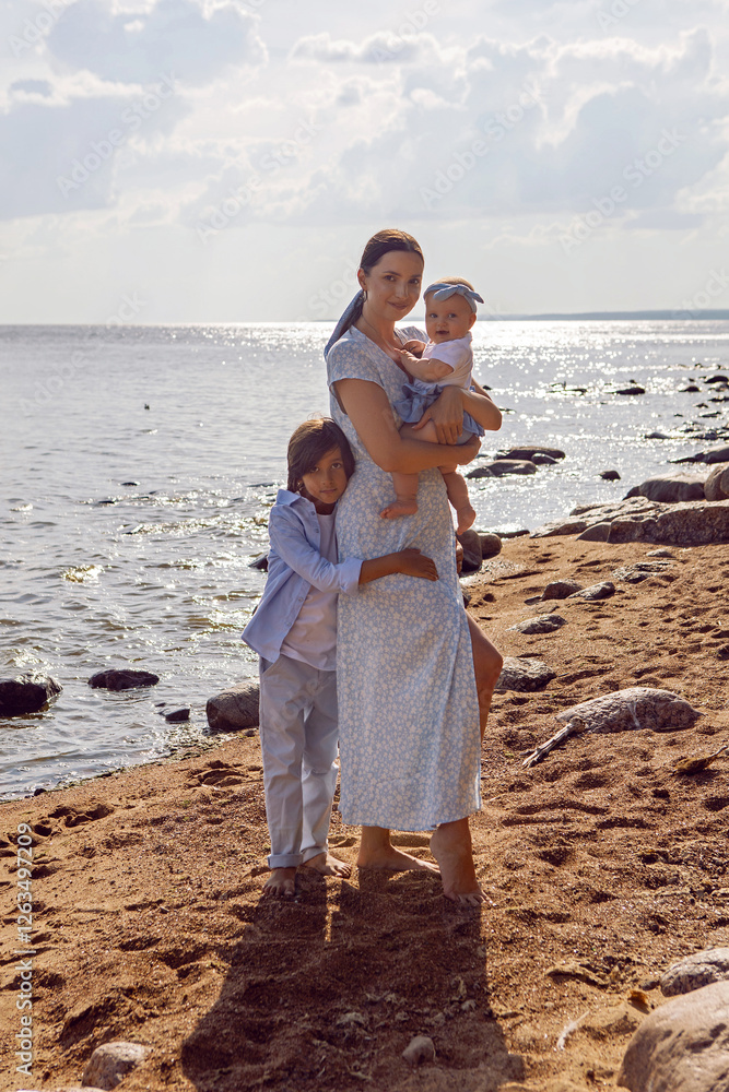 A happy mother in a dress with two children aged seven and a baby are standing on a sandy beach by the sea in summer