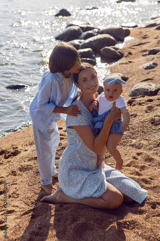 A happy mother in a dress with two children aged seven and a baby are standing on a sandy beach by the sea in summer