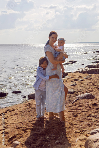 Obraz na płótnie A happy mother in a dress with two children aged seven and a baby are standing on a sandy beach by the sea in summer w salonie
