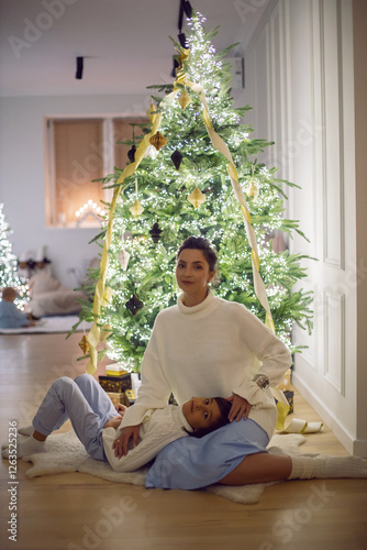 A mother in a white sweater sits with her son for 7 year on the floor by the Christmas tree in an apartment on New Year's Eve