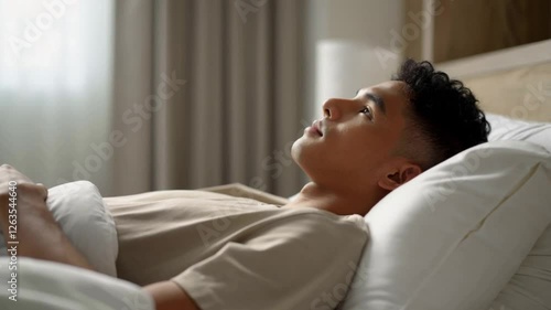 A young man is lying in bed, thinking about something important. His gaze is directed into the void, and the atmosphere of the room creates a feeling of coziness, calmness and light thoughtfulness.