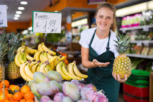 Fotografie Smiling fifteen-year-old girl who works part-time in a store as a trainee saleswoman demonstrates a pineapple, holding it
